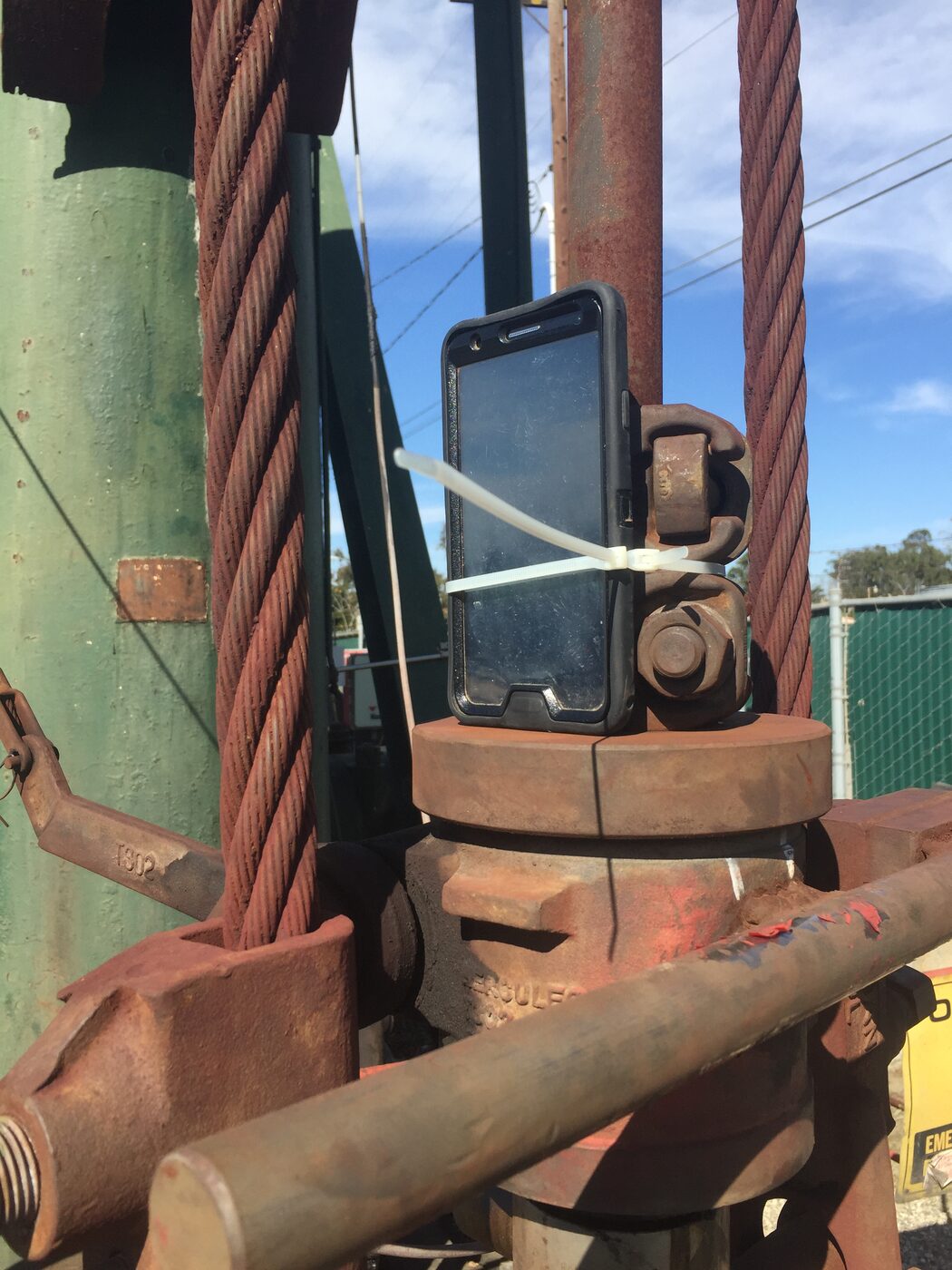 Photo: a smartphone in a black case zip-tied to the rusty rod rotator clamp of a pumping unit, with the bridle's wire ropes, green-painted equipment, and blue sky behind it.